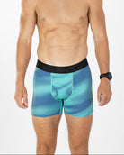 A man modeling blue and teal patterned boxer briefs with a black waistband, photographed against a clean white studio background.