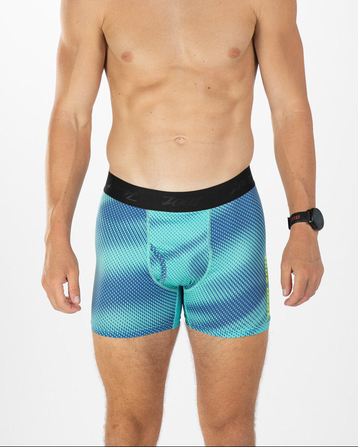 A man modeling blue and teal patterned boxer briefs with a black waistband, photographed against a clean white studio background.