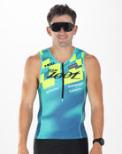 A man wearing a sleeveless cycling jersey with a blue and yellow gradient pattern, black cycling shorts, and black sunglasses, standing against a white studio background.