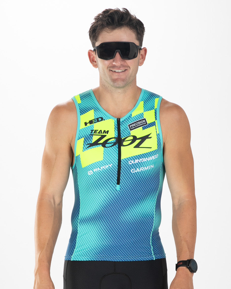 A man wearing a sleeveless cycling jersey with a blue and yellow gradient pattern, black cycling shorts, and black sunglasses, standing against a white studio background.